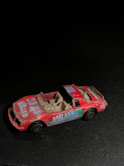 A close-up, high-angle shot shows a damaged, red toy race car lying on its side against a black, textured background. The car is worn and appears to be made of metal. There is visible dirt and wear on the car's surface, indicating age and use. The car has "33" prominently displayed in blue lettering on its side, along with the text "MR. RED". Some blue lettering is also visible on the car's rear fender, reading "AIR RED". The car's wheels are black and appear to be intact, though one is slightly tilted. The interior of the car, visible through a gaping hole in the roof, is a light beige color. The overall scene suggests an indoor setting, possibly a child's room or a storage area, with poor lighting. The specific location of Yelizovo, Russia, provides geographical context but no direct visual cues within the image itself. No people are visible, and there are no indications of time of day or weather. The image is focused on the solitary, damaged toy.