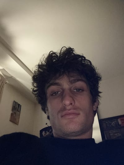A male individual is depicted in an indoor setting, reportedly in Vincennes, France. The subject, a young adult, occupies the majority of the frame, facing forward with a direct gaze toward the camera. He has dark, curly hair and light-colored eyes. Minor stubble is visible on his chin and jawline. He is wearing a dark, high-neck garment. Overhead artificial illumination highlights the subject's features and the light-colored ceiling. The background on the left reveals a light-toned wall with a partial view of a light-colored curtain and a rectangular, framed item. The background on the right contains darker, indistinct elements, including a vertical boundary and a faint reflection showing dark, unidentifiable shapes.