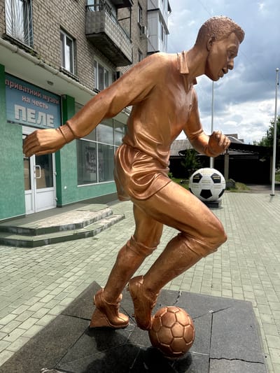 In the city of Lopaskyne, Ukraine, a bronze statue of a football player, likely representing Pele, stands in motion on a paved area. The statue depicts the player with his right leg forward, poised to kick a football at his left foot. A large, black and white football is situated behind the statue. The background features a building with windows and a sign that reads "приватний музей на честь ПЕЛЕ" (Private Museum in Honor of Pele), indicating the location is a museum dedicated to the famous footballer. The sky above is cloudy.