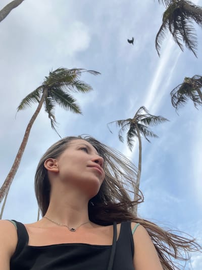 A low-angle, medium shot captures a woman from the chest up, looking towards the upper right of the frame. Her face is in profile, with her head tilted slightly upward and her gaze directed away from the camera. Her hair, a mix of brown and lighter streaks, is windblown, framing her face. She wears a simple black tank top and a delicate silver necklace with a small cloud-shaped pendant.
Behind her, the sky is a mix of pale blue and white clouds, with the tops of several tall palm trees reaching upwards. One palm tree on the left is particularly prominent, its trunk textured and its fronds spread out. Another palm tree is visible in the upper right corner, and a third is positioned further back. A small bird in silhouette is captured in flight against the cloudy sky in the upper middle portion of the image. The overall impression is one of a breezy day outdoors, possibly near a tropical coast, with the natural elements of sky, trees, and wind playing a significant role.
The image is taken in Thalaramba, Sri Lanka.