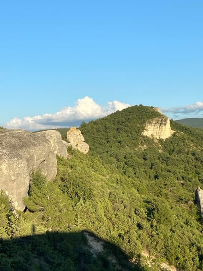 A wide shot captures a natural landscape with a prominent rock formation and a forested mountain under a clear blue sky. In the foreground, large, weathered grey boulders dominate the left side of the frame, casting shadows across a dense, green forest that covers the sloping terrain. To the right of the boulders, a smaller, lighter-colored rock formation is visible, appearing somewhat weathered and rounded.
Further in the background, a mountain rises, densely covered with green trees. A significant portion of the mountain face is a sheer, light-colored cliff, revealing stratified rock layers. White, fluffy clouds are scattered across the upper part of the sky, partially obscuring the distant horizon line. The scene appears to be a natural, undeveloped area, likely a mountainous or hilly region. The specific location is identified as Krasnomakskoye Rural Settlement, Ukraine.