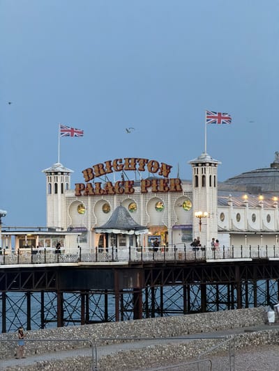 The image shows a British pier under a clear sky, likely taken during the day or early evening. The pier, constructed with white painted structures and intricate metalwork, extends over the water. A large sign with red lettering that reads "BRIGHTON PIER" is prominently displayed. Two Union Jack flags are visible, one on a flagpole near the entrance and another further down the pier, flapping in the breeze. Several people are scattered along the pier, some walking and others sitting, enjoying the atmosphere. Small businesses, possibly souvenir shops or amusements, are visible along the pier's edge, indicated by signs such as "GIFT SETS" and "COFFEE ICE CREAM PIES." The scene conveys a sense of leisure and vacation. The water below the pier is a deep blue, reflecting the sky. The overall impression is a classic seaside attraction on a pleasant day.