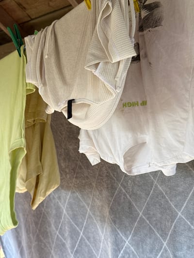 A close-up, outdoor shot shows several pieces of clothing hanging on a clothesline. The garments include a light green ribbed t-shirt on the far left, a light yellow t-shirt next to it, and a cream-colored ribbed garment layered over a white t-shirt with green text. The clothesline is made of wood and is supported by wooden beams. The background consists of a grey, diamond-patterned screen. The scene appears to be in a backyard or open area, likely in a residential setting. No individuals or specific landmarks are visible. The overall impression is of laundry being air-dried. It is not possible to determine the exact location in Ivatsevichy, Belarus from the provided image.