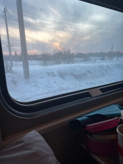 This image captures a winter landscape viewed from inside a moving train in Pervomaiskoe, Russia. The scene outside the dark-framed train window displays extensive snow cover across uneven terrain. Utility poles with electrical wires traverse the midground, accompanied by sparse, bare trees and the silhouettes of distant buildings against the horizon. The sky above is overcast with grey clouds, from which a bright, localized yellow-orange light, indicative of a sunrise or sunset, emerges towards the upper-left, illuminating the cloud layers. Inside the train, a light brown window sill is visible, holding a small, light-colored card tucked into the frame. Below the sill, on an interior shelf, a teal rolled item is situated next to several stacked pink circular containers and a partially visible white disposable cup with a red band. A white fabric object, possibly a pillow or blanket, is partially visible in the bottom-left corner of the frame.