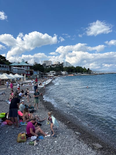 The image shows a crowded pebble beach on a sunny day. People are scattered across the beach, some sitting on towels and blankets, others standing or walking. A few individuals are in the water. In the background, there are buildings and trees along the coastline. The sea is blue with gentle waves. The scene is set in Alushta, Ukraine.