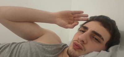 A person is pictured from a low angle. He faces the camera and forms an "OK" gesture with his right hand. His head is tilted to the left, and he has short brown hair and dark eyebrows. He is wearing a gray t-shirt and a white garment near his shoulder. The background is a plain white wall, and a necklace is visible around his neck.