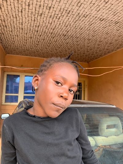 In this low-angle, portrait-style, rotating video, a young person with braided hair and a gold hoop earring is looking up at the camera. The subject is wearing a black long-sleeved shirt. Their skin appears smooth and luminous. The background is a brick wall with a reddish-brown hue. To the right of the subject, a portion of a dark-colored vehicle is visible, reflecting the sky and surrounding environment. A window with blue panes is also present in the background, suggesting an outdoor setting or a space with an open-air feel. The lighting suggests it is daytime, and the overall mood is casual and relaxed. The image appears to be taken in Barakin Sabawa, Nigeria.