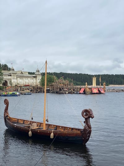 The image shows a body of water with a Viking longship in the foreground. The ship is made of wood and has a carved ram's head at the bow. In the background, there is a large, fortified building resembling a castle, with towers and flags. There are also several other wooden structures and boats docked at a pier. Beyond the structures, a dense forest can be seen, and the sky is overcast. The scene appears to be a historical reenactment or a themed attraction, likely located in a waterfront area. The specific location is the "Vikings" park in Motornoe, Russia, which is known for its authentic reconstructions of Viking settlements and ships.