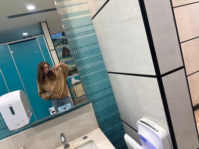 The image depicts a public restroom in Nong Prue Subdistrict, Thailand. A young woman with curly brown hair is taking a mirror selfie. She is wearing a tan sweatshirt and blue jeans. She is holding a silver smartphone in her right hand, with her left hand on her hip. To her left is a white automatic soap dispenser with the brand name "SVAVO" visible. Behind her, in the mirror's reflection, are bright blue bathroom stall doors with ornate white patterns. Further into the reflection, a large mirror displays a landscape image of a rocky island with trees and a body of water. The restroom walls are tiled with a gradient of light blue and white mosaic tiles, transitioning to large white marble-patterned tiles on the right side of the frame. A white automatic hand dryer with the brand name "NEOMATE" is visible on the lower right side of the image, emitting a blue light. A silver faucet is present above a cream-colored sink. A sign with Thai text and "TISSUE PAPER HERE" in English is affixed to the mirror.