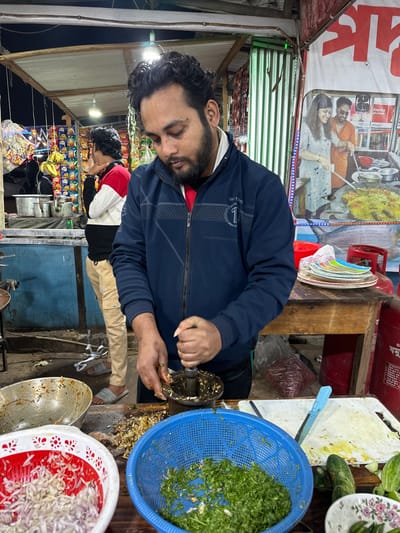 A man in a blue jacket is preparing food at a stall. He is operating a food grinder next to a blue basket filled with chopped greens. A red and white basket of chopped onions sits nearby, along with dirty dishes. Another man in a red and white jacket stands near shelves stocked with snack foods. A food advertisement hangs above the stall.