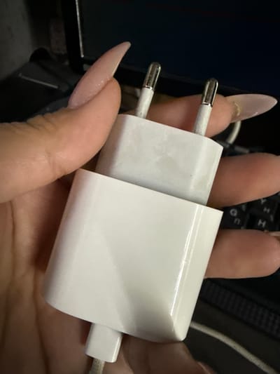 The image shows a close-up of a white power adapter with two metallic prongs being held by a person's hand. The adapter is partially obscured by the hand, which has long, light-colored artificial nails. The background is dark and appears to be an indoor setting, possibly near a keyboard. There is no discernible location in the city of Taldykorgan, Kazakhstan.