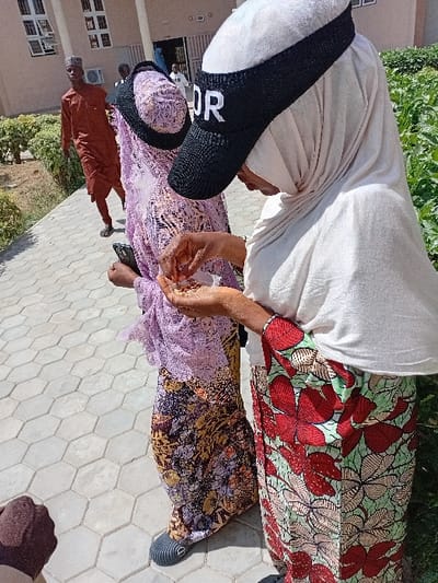 The scene depicts an outdoor setting in Kwazailewa, Nigeria, under bright daylight. In the immediate foreground, a female subject wears a light-colored hijab and a black visor with "DOR" in white uppercase letters across the front. She holds a small quantity of light-colored granular material in her cupped hands. Her lower attire is a patterned fabric featuring large red floral designs on a light green background. Adjacent and slightly behind her, a second female subject wears a purple patterned head covering and a multi-colored patterned wrapper. She holds a dark smartphone in her left hand. Her footwear consists of dark open-toed sandals on a hexagonal paved walkway. In the midground, a male subject walks away from the camera, looking over his left shoulder towards the foreground. He wears a reddish-brown traditional tunic and trousers, complemented by a dark cap. The background features a light-colored building with multiple rectangular windows and an open entrance, with green foliage visible to the right. The ground is consistently paved with light grey hexagonal tiles.