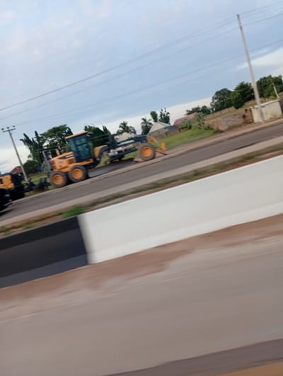 This image captures a daytime scene in Uke, Nigeria, depicting heavy construction equipment parked alongside a road. The perspective is from a moving vehicle, evidenced by motion blur in the foreground and a slight tilt to the right.
The primary subject is a large yellow motor grader, positioned on the shoulder of an asphalt road. This grader features large orange-brown wheels, a visible driver's cabin with dark windows, and a long front-mounted blade. To the left of the grader, partially obscured, is another yellow and black piece of heavy machinery, possibly an excavator or loader, also stationary.
The road itself appears to be recently laid or under construction, with a dark asphalt surface. An unpaved, lighter-colored shoulder runs alongside the asphalt. In the immediate foreground, a white concrete barrier with a dark top edge runs diagonally across the bottom half of the frame, separating the viewer's path from the main road and construction site.
In the background, behind the construction equipment, a line of various trees and green foliage is visible. Among the trees, several residential-style buildings with light-colored walls and dark roofs are discernible. At least two utility poles, strung with multiple horizontal power lines, are present on the left and right sides of the background, extending above the trees. The sky is overcast with a light blue-grey hue, indicating cloudy conditions. No individuals are visible in the frame.