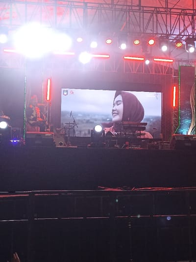This media file depicts a lively outdoor stage event taking place at night in Kecamatan Pujut, Indonesia. The central feature is a large LED screen displaying a close-up image of a young woman smiling joyfully, wearing a dark head covering (hijab). The background behind her on the screen appears to be an open, possibly natural or rural, landscape.
The stage is structured with an elaborate metal truss system overhead, from which numerous bright stage lights, in white and red hues, illuminate the area. These lights create a dynamic atmosphere, with some causing noticeable lens flare in the image. To the left of the main screen, a person, possibly a performer or crew member, is partially visible, obscured by bright lights, and appears to be wearing distinct attire.
Musical instruments are arranged on the stage, including a drum kit, a keyboard on a stand, and a microphone stand. Faintly visible text on the bass drum seems to read "Go Drum". On the top left of the LED screen, a logo featuring a stylized "Go" is present, and some partially obscured text, possibly "PONTIA", can be seen near the bottom of the screen.
The foreground is dark, revealing the top of a barrier or railing, hinting at an audience beyond. The overall setting, with its bright lights, musical equipment, and large display screen, suggests a concert, festival, or similar public performance or event. The time of day is clearly nighttime, enhancing the dramatic effect of the stage lighting.
