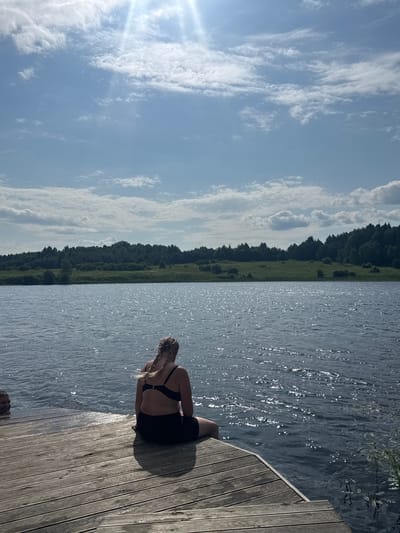 A medium shot captures a person with blonde braided hair, facing away from the camera, seated on a wooden dock. They are wearing a black bikini top and black shorts, with their legs dangling over the edge of the dock. The dock extends towards a body of water that reflects the sunlight, creating a shimmering effect. Across the water, a lush green landscape with a dense line of trees forms the background, under a bright blue sky dotted with white clouds. The scene appears to be set in Pozdyshki, Russia, on a sunny day.