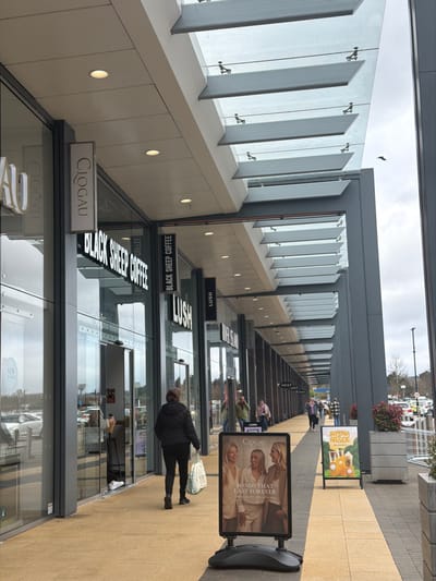 The image depicts an outdoor retail precinct in Bretton, United Kingdom, under an overcast sky. A modern building features a continuous facade of large glass storefronts and automatic sliding doors, covered by an extended overhead canopy. This canopy comprises transparent glass panels set within a grey metal framework, incorporating horizontal louver-like elements and recessed ceiling lights.
Prominent store signage includes 'Clogau' (also seen on a vertical banner sign), 'BLACK SHEEP COFFEE,' 'LUSH,' 'RIVER ISLAND,' and 'NEXT.' The paved walkway consists of tan and light brown tiles. A woman, wearing a dark jacket and dark trousers, walks away from the camera along this pavement, carrying a light-colored shopping bag in her left hand. A person is partially visible inside the 'BLACK SHEEP COFFEE' store through its glass doors. Further along the walkway, several other individuals are observed walking or standing.
Two A-frame signs are positioned on the pavement: one for 'Clogau,' displaying three women and the text "BONDS THAT LAST FOREVER," and another, located further from the camera, advertising 'Banana Arcade' with illustrations of various beverages. Concrete planters containing red-leafed shrubs are placed periodically. Beyond the retail establishments, a parking lot with numerous parked vehicles and lampposts is visible. A solitary bird is in flight in the upper right portion of the sky.