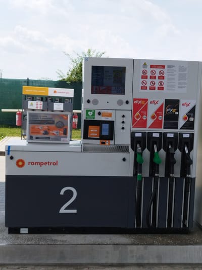 This is a full-frame, eye-level, outdoor, medium-shot photograph of a Rompetrol gas station in Clinceni, Romania. The gas pump, identified by the large numeral "2" on its side, is the primary subject. The pump has four dispensing nozzles, two of which are green and two are black. Above the nozzles, on the right side of the pump, there are several placards with logos and product names such as "efixS", "Benzina 98 E10", "Benzina 95 E10", "Diesel 55 B7", and "Motorina 51 B7". On the left side of the pump, a digital display shows "130,11" and "14,99" next to currency symbols. There is also a framed advertisement with Romanian text, displaying various offers such as "30x", "10x", and "2x". Two red fire extinguishers are visible mounted on the side of the pump station. In the background, a green fence runs horizontally across the frame, with some trees and a cloudy sky above. The gas pump is situated on a concrete surface.