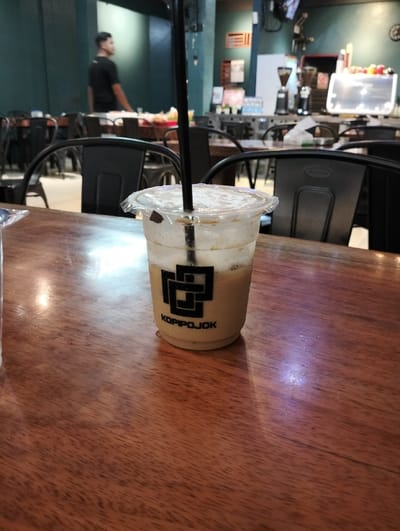 The image displays the interior of a coffee shop identified by the "KOPIPOJOK" logo, located in Jeunieb, Indonesia. In the foreground, a transparent plastic cup containing a partially consumed iced coffee with a black straw and a clear lid rests on a polished wooden table. The cup exhibits condensation and features the "KOPIPOJOK" logo in black text. The midground contains multiple dark-colored, industrial-style metal chairs and wooden tables. In the background, a male individual wearing a black t-shirt is visible standing behind a table, partially obscured and facing right. Beyond the seating area, a service counter is equipped with professional coffee machinery, including a silver espresso machine with multi-colored nozzles and two coffee grinders. The interior walls are painted a dark teal or green color, with various signs or decorative elements affixed.