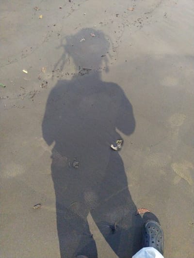 The image depicts a sand surface, likely near a beach or riverbank. The shadow of a person is cast on the sand, revealing the presence of a person with what appears to be dreadlocks and a casual pose. To the right, a person's foot wearing a Croc-style shoe is visible, with part of a white material near it. Various small pieces of organic debris, like leaves and twigs, are scattered on the sand. No explicit information about the location of Badem, India, can be determined from the image.