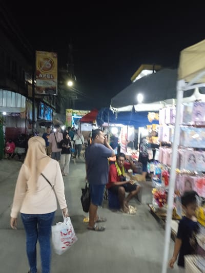 The image depicts a street scene in Kauman, Indonesia, at night. Several people are walking along a street lined with stalls selling various goods. One person is wearing a light-colored headscarf and carrying a white shopping bag. A man is standing nearby with his hand to his head, wearing a blue shirt and denim shorts. A seated man is wearing a red shirt and a yellow scarf around his neck. A child is standing near a stall with various colorful items on display. In the background, there is a sign with the word "APEL" and a logo. The stalls are illuminated by overhead lights. The street appears to be crowded.