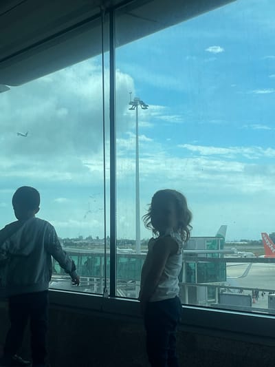 The image, correctly oriented, shows two young children standing by a large window, looking out onto an airport tarmac. A young girl, with curly hair and wearing a light top and dark trousers, is positioned on the right side of the frame, gazing intently outwards. To her left, a young boy, wearing a patterned top and dark trousers, is also looking out and has his right arm extended, pointing towards the scene outside.

The children are primarily silhouetted against the bright light from outside, making their facial features indistinct but highlighting their posture of observation. Outside the window, under a bright, cloudy sky, an airport apron is visible. An easyJet airplane, identifiable by its orange tail fin and "easyJet" logo, is parked on the tarmac. Various airport structures, possibly jet bridges or other terminal buildings, are also visible. The scene captures a moment of interest and wonder for the children as they observe the activity at what is identified as Moreira, Portugal, during the daytime with a bright, overcast sky.