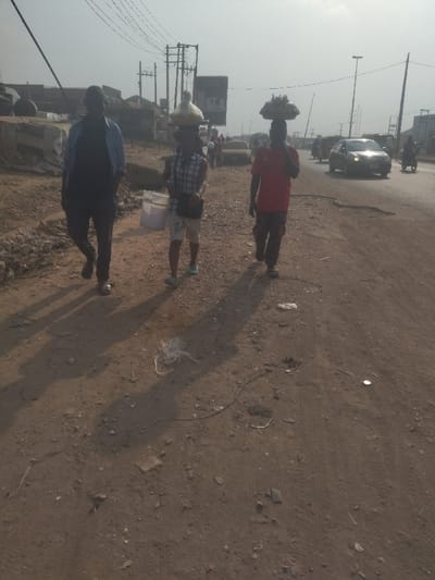 The image shows three people walking along a dirt road in Way, Nigeria. The people are walking in the same direction, towards the camera. Two of the people are carrying items on their heads: one is carrying a large bundle of white material, the other is carrying a large bundle of food items. The third person is carrying a white bucket. The road is dusty and brown, with some small rocks and debris scattered along the side. The environment is dusty and hazy. There are buildings and a few trees visible in the background. The road is a busy thoroughfare, with several cars, motorcycles, and pedestrians traveling in both directions. The road is located in the city of Way, Nigeria, at the coordinates 9.029 latitude and 7.584 longitude. The sky is clear and blue with a few wispy clouds.