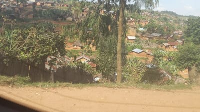 A hillside village is depicted, with simple houses densely packed together. The houses feature a mix of thatched and corrugated metal roofs, and their walls are predominantly made of reddish-brown material. Green vegetation, including trees and shrubs, surrounds the village. A dirt road runs along the foreground, and a wooden fence is visible near the roadside. The overall scene suggests a rural and somewhat impoverished setting.