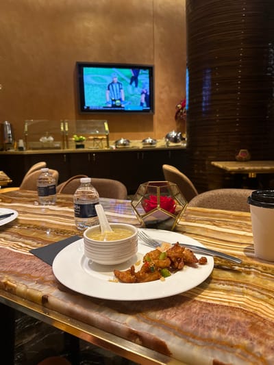 This image captures a well-lit indoor dining or lounge area within an MGM Resorts International property located in Paradise, Nevada, United States. In the foreground, a polished, variegated brown, beige, and white natural stone table holds a meal consisting of a white ceramic plate with several pieces of golden-brown fried food garnished with chopped green and red ingredients, a small white ceramic bowl containing light-yellow soup with a matching spoon, a silver fork and knife, and a black napkin. Two clear plastic water bottles, one labeled "MGM Rewards," are present alongside a geometric gold-framed glass object containing red roses and a white disposable cup with a black lid.
In the background, a flat-screen television mounted on a warm brown wall displays an American football game, featuring a referee in a striped uniform. Below the television, a dark counter serves as a buffet line, equipped with multiple silver chafing dishes, two gold-framed glass display cases, and a silver beverage dispenser. Brown upholstered chairs are visible around tables. A large, dark brown, textured cylindrical pillar stands on the right side of the frame. No individuals are present in the immediate view.