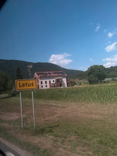 This is a photograph of a rural scene in the village of Letuš, which is part of the municipality of Šmartno ob Paki in Slovenia. A yellow sign with black lettering that reads "Letuš" is prominently displayed in the foreground, supported by two metal poles. Behind the sign, a modern house with a red roof and solar panels is visible. The house is situated amidst a field of tall green crops, likely corn. In the background, a densely forested hill rises under a bright blue sky with scattered white clouds. The overall environment suggests a sunny day in a countryside setting.