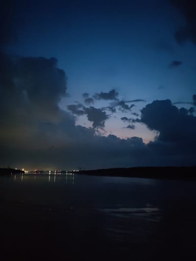 The image depicts a twilight scene over a body of water, likely a river or a bay, in the city of Lapach, Russia. Dark, dramatic clouds dominate the upper portion of the frame, obscuring much of the sky and creating a somber atmosphere. A faint band of light is visible near the horizon, suggesting the last vestiges of daylight or artificial illumination from a distant cityscape.

On the far bank of the water, a faint line of lights indicates the presence of a town or industrial area. These lights are reflected in the calm water, creating streaks of white and yellow against the dark surface. The foreground is entirely dark, with the water appearing black and reflective. The silhouette of trees or dense vegetation can be seen along the far bank.

No people or specific actions are discernible in the image. The overall impression is one of stillness and quiet, with nature's elements, the dark clouds and water, taking precedence. The scene is likely captured from a vantage point near the water's edge, possibly a pier or a riverbank. The low light conditions and the dark palette of blues and blacks suggest this is either dawn or dusk.