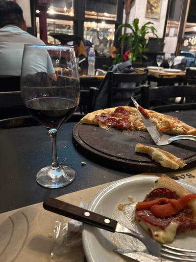 The image displays an indoor scene within a pizzeria, likely *El Tio*, in Mar del Plata, Argentina, during the evening. In the immediate foreground on a black table surface, a stemmed wine glass, approximately three-quarters full of red wine, is positioned. Adjacent to it, a white ceramic plate holds a partially consumed slice of pizza topped with pepperoni, melted cheese, and strips of red bell pepper. A metal fork and a knife with a dark wooden handle and two silver rivets rest on this plate.
In the midground, a circular dark wooden pizza board contains two remaining large slices of pizza; one half features pepperoni, while the other has a lighter-colored, chunky topping. A metal pizza spatula, with a thin red accent on its handle, lies across the pizza, and a discarded piece of pizza crust is visible on the board.
The background reveals a restaurant interior with dark wooden tables and chairs. A person wearing a light blue striped short-sleeved shirt is seated with their back to the camera, partially visible on the left. Through large windows, a blurred exterior street scene is discernible, including reflections of interior lights and a distant sign. The restaurant is decorated with a tall green potted plant, a smaller plant with red flowers, a blue star ornament, and a metallic rooster figurine. The setting is illuminated by artificial lighting.