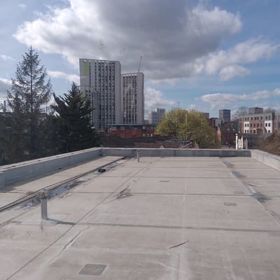 The image shows a cityscape in Coventry, United Kingdom, viewed from a flat, concrete roof. In the background are several tall, modern buildings, including one with "CODE" vertically displayed on its side. A red crane is visible behind the buildings. In front of the buildings are trees, residential houses, and other urban structures. The sky is partly cloudy with a mix of white and dark clouds. The foreground is dominated by the flat, grey roof with rectangular sections and some visible drainage or utility elements. The roof itself appears weathered with slight discoloration and minor debris. There are no people visible in the scene.
