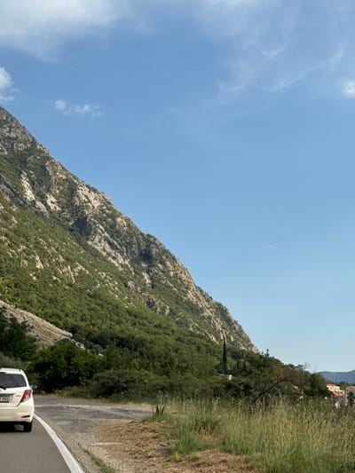 A white Toyota Yaris hatchback is parked on the side of a road that curves to the left, with its rear facing the camera. The vehicle is positioned on the asphalt, next to a white line marking the edge of the road. The road itself is lined with dry grass and vegetation.

To the left of the road, a steep, rocky mountainside rises sharply, covered in dense green trees and shrubbery. The mountain's terrain is rugged, with visible rock formations and a scattering of lighter-colored stone. The sky above is a clear, bright blue with a few wispy white clouds.

In the distance, nestled among the trees on the lower slopes of the mountain, are several buildings with red-tiled roofs, suggesting a small settlement or village. The overall environment appears to be a rural or semi-rural area, likely in the region of Donji Orahovac, Montenegro, given the characteristic mountainous landscape and coastal vegetation. The scene depicts a roadside view with a focus on the natural environment and the presence of a vehicle.