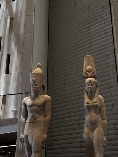 The image shows two large, ancient Egyptian statues within a modern building. The statue on the left depicts a male figure wearing a headdress, a kilt, and a broad collar. The statue on the right appears to be a female figure, also wearing a headdress and jewelry. The statues are made of a speckled stone and have a weathered appearance. They are positioned in front of a wall with a textured, segmented design. To the left of the male statue, there is a contemporary building facade with vertical and horizontal elements, and two visible security cameras. The lighting is dim, with some areas in shadow and others illuminated, creating a dramatic effect. There is no indication of the city of Kafrat Nassar, Egypt.