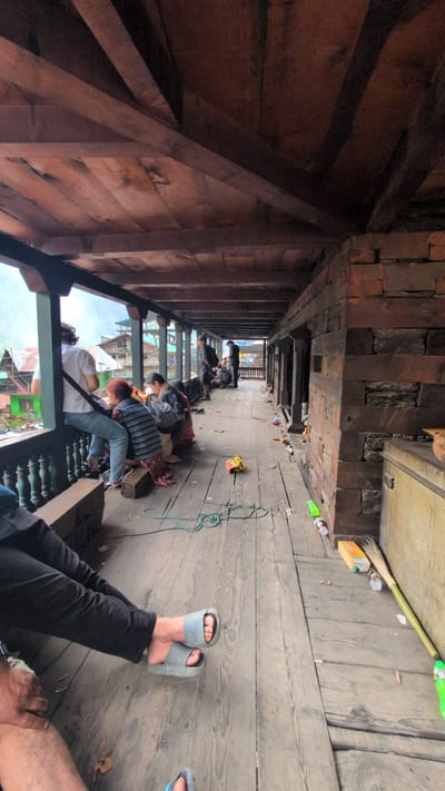 A group of people are gathered on a wooden balcony in Malana, India. The balcony is covered by a wooden roof supported by exposed beams. The floor is made of weathered wooden planks. To the left, several individuals are seated or leaning against a railing, looking out at a view of buildings and greenery. To the right, the balcony opens onto a hallway with more people standing and sitting. The floor is littered with various objects, including plastic bottles, wrappers, and a coiled green rope. A broom and a closed wooden chest are also visible on the right side. The walls are constructed of stone. The overall impression is of a rustic, communal space.