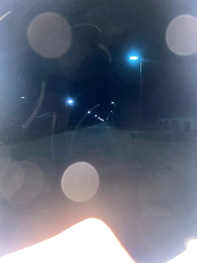 The image depicts a nighttime street scene, presumed to be located in Aigiere, Nigeria. The photograph was captured from a low vantage point, likely through a reflective or partially obscured surface, as indicated by numerous circular light flares and linear streaks across the frame.

A straight, unidentifiable road surface, appearing light grey, extends centrally into the distance, flanked by streetlights. Multiple tall streetlights with bright, blue-white light sources illuminate the road, receding in a linear pattern. A single prominent streetlight is visible on the right side of the road in the mid-ground. On the right side of the scene, in the middle distance, a light-colored building with multiple arched openings on its facade is discernible. Faint structural outlines are also visible on the left side of the road. No individuals, animals, or additional vehicles are present in the observable environment. A strong, overexposed light source occupies the lower portion of the image, causing significant glare. The sky is dark, consistent with nighttime conditions.