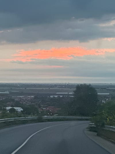 The image captures a serene landscape scene at either sunrise or sunset, viewed from an elevated position. The sky occupies the left side of the frame (if rotated to a standard landscape orientation), featuring a dramatic display of clouds. A prominent band of clouds is intensely illuminated with vibrant orange and pink hues, indicating the presence of the sun low on the horizon, while the surrounding sky is a softer grey-blue.

Stretching across the middle of the image is a hazy horizon line, revealing a distant landscape. This area appears to be a valley or plain, dotted with indistinct buildings and clusters of trees, suggesting a rural or semi-urban settlement. The lighting across this distant land is subdued, adding to the atmospheric quality of dusk or dawn.

On the right side of the image, a paved road or pathway dominates the foreground, curving upwards and to the right. A clear white line marks the edge of the road. Along the immediate edge of the road and extending into the lower right corner, there is dense, dark green foliage, likely consisting of trees and bushes. Another segment of dark green trees is visible further down the slope, separating the road from the more distant landscape.

No people or specific activities are visible in the image. The scene conveys a sense of tranquility and natural beauty, characteristic of a scenic overlook. There is no visible text. The location context indicates this is a landscape in Isperihovo, Bulgaria.
