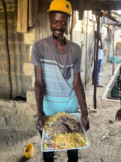 A man wearing a yellow New York Yankees hat and a striped shirt is holding a white tray of food. The tray contains what appears to be beans and a yellow-colored vegetable mixture. The man is standing in an open-air space, possibly in a courtyard. There is a second individual, partially obscured, wearing a white shirt and black pants, who appears to be standing in the background. The location is Githurai, Kenya, based on the coordinates -1.204 latitude and 36.905 longitude. The scene suggests a casual, everyday interaction between the two individuals, possibly a meal shared between friends or family. The environment appears to be a simple, outdoor area, possibly in a residential neighborhood. There are wooden beams in the background, possibly indicating a partially constructed or unfinished building. The image is captured in a slightly low angle, which provides a close-up view of the man and the food. The lighting is natural, suggesting a daytime setting.