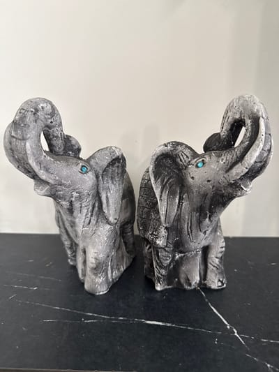 Two grey elephant figurines with upturned trunks and small turquoise eyes are positioned side-by-side on a dark, reflective surface, possibly a countertop or shelf. The figurines appear to be made of stone or a similar textured material, with visible lines and imperfections suggesting a handcrafted quality. The background is a plain, light-colored wall. The scene is indoors. The provided image does not contain information to determine its location in Khokhryaki, Russia.