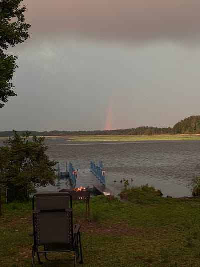 An outdoor scene depicts a tranquil lake with a light rain falling, indicated by ripples on the water's surface. In the foreground, a dark gray folding chair sits on a grassy area. To the right of the chair, a metal barbecue grill contains glowing embers. Beyond the grill and chair, a blue pier extends into the water. A small group of ducks swims near the pier. Across the lake, a dense forest line borders a strip of marshland, and a faint rainbow is visible in the overcast sky. The scene suggests a natural setting within the Слабодкаўскі сельскі Савет, Belarus.