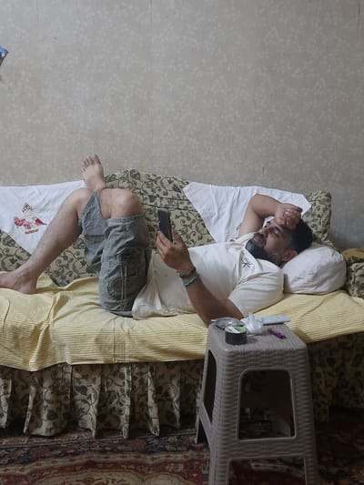 An adult male is observed reclining on a couch or bed in an indoor residential setting, located in Cairo, Egypt. He has a dark beard and mustache, and is wearing a cream-colored t-shirt with a small "CK" logo on the left chest, and grey cargo shorts with a black star-shaped patch on the right thigh pocket. His feet are bare. He is holding a black smartphone in his right hand, while his left hand is resting on his forehead or covering his eyes. Bracelets, one blue and one dark, are visible on his left wrist.
The couch or bed is covered with a yellow and white striped sheet, beneath which is a base featuring a dark green floral/leaf pattern on a beige background. A white pillow supports his head, and a white decorative cloth embroidered with red birds and butterflies is draped over the head of the couch/bed.
To the right of the man, a light beige/brown plastic stool with a woven texture serves as a side table. On its top surface are a small black cylindrical container, a crumpled white plastic bag or wrapper, a small white rectangular item, and a purple pen or marker.
The wall behind the couch is covered in light beige wallpaper with a subtle, lighter-toned floral or leaf pattern. The floor is covered by a patterned rug featuring shades of red, blue, and beige in an intricate design. The scene depicts a moment of rest or contemplation within a domestic interior.