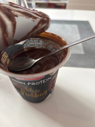 A close-up, slightly angled shot shows an open container of chocolate pudding with a spoon inside. The pudding is dark brown and appears smooth, with swirls from being stirred. The container is black with text in gold and white that reads "HIGH PROTEIN Choc Pudding." Part of the lid, also with chocolate residue, is visible in the upper left. The pudding is on a white surface, possibly a table, with a grey placemat underneath. The background is blurred but suggests an indoor environment with shelves and possibly kitchen appliances. The scene is not specific to Groß-Gerau, Germany, or any other city.