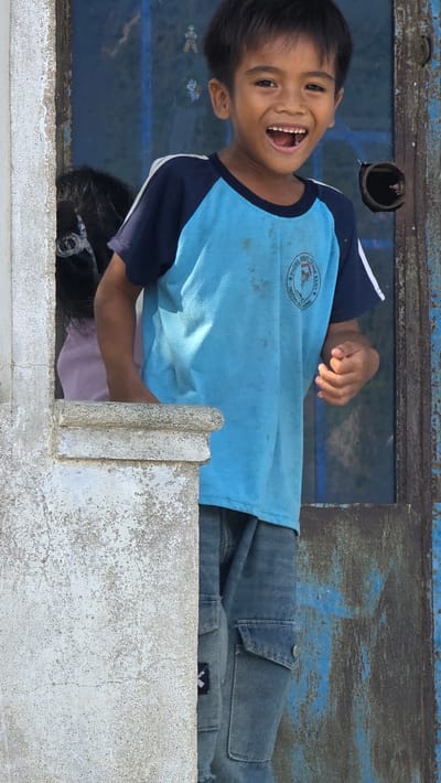 A boy in a blue t-shirt is smiling broadly and appears to be positioned inside of a structure in Ba Ngoi Ward, Vietnam. The boy's dark hair is short, and he is wearing a shirt with dark blue sleeves and a logo on the front. He is also wearing jeans. The structure he is in has a window with what appears to be painted designs on it. Behind him is some rusted metal that is blue in color. In front of the boy is a concrete surface. Another person, possibly a child wearing glasses, is partially visible behind the concrete structure near the boy.