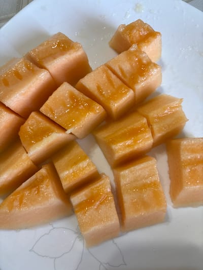 A white plate holds several cut pieces of cantaloupe melon, arranged in rows. The melon pieces are orange and appear to be drizzled with a light-colored syrup or glaze. The plate has a subtle floral pattern on its surface. The lighting suggests an indoor setting. This image was captured in Cheshnegirovo, Bulgaria.