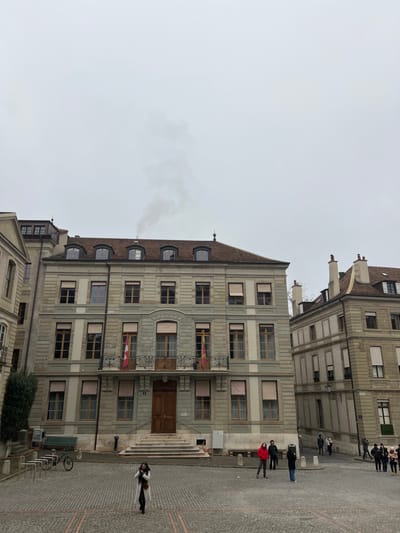 This media file captures an outdoor scene in what appears to be a square in Geneva, Switzerland, on an overcast day. The central focus is a multi-story building with classical architecture, featuring a light grey stone facade, numerous windows, and a dark, sloped tiled roof. Several windows on the main building have beige awnings, some extended, and small red flags with white crosses (likely Swiss flags) are displayed from a few balconies and windows on the first floor. A prominent wooden double door serves as the main entrance, accessible via a set of stone steps. Above the entrance, a balcony with ornate iron railings extends across the first floor. On the roof, dormer windows are visible, and smoke rises from a chimney on the left side.
To the right and slightly behind the main building, another similar stone building can be seen. The ground is paved with cobblestones, forming a public square. Several people are scattered across the square; one person in a white coat walks away from the main building, while a small group, including an individual in a red jacket, stands near the building's steps. Further back, more individuals and groups are visible. A green park bench and a bicycle parked at bicycle racks are present on the cobblestones to the left of the main entrance steps. The sky is uniformly grey and overcast, suggesting a cool, cloudy daytime. No discernible text is visible in the image.
