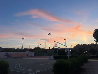 The image captures an outdoor basketball court and its adjacent environment at dusk or dawn in Picassent, Spain. The upper portion of the frame is dominated by a clear sky exhibiting a gradient from deep blue overhead to soft pink, orange, and pale yellow tones near the horizon, accented by wispy, horizontally stretched clouds reflecting these colors. The primary subject is a dark green basketball court outlined with white markings, including free-throw lines, the key, and a half-court circle. On the right, a basketball hoop with a transparent backboard and a white support structure is visible. Multiple tall, dark poles topped with floodlights are positioned across the scene, suggesting nighttime usability for the sports area; one prominent pole stands in the mid-ground. Along the left side of the court, a light-colored wall features faded murals depicting basketballs and abstract patterns, located behind a green fence. Beyond this, another court, possibly for tennis or padel, with a reddish surface, is partially visible. Dark green bushes and trees frame the court and foreground. In the background, several multi-story residential buildings, characterized by numerous windows and rooftop structures like antennas and satellite dishes, are silhouetted against the horizon. A concrete structure, potentially a trash receptacle or seating, is situated near the basketball hoop. No individuals are present.