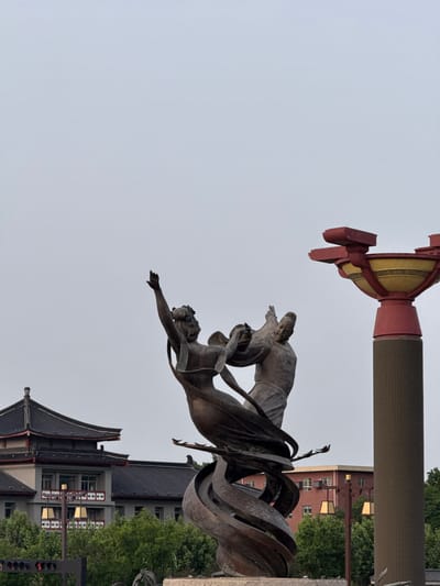 A low-angle shot depicts a bronze statue in Lintong District, China. The statue features two figures enveloped in flowing ribbons, set against a backdrop of a traditional Chinese building with a dark roof and light-colored walls. A red and gold architectural structure stands adjacent to the statue, and the sky is overcast. In the background, glimpses of modern buildings and green foliage are visible. A traffic light pole is present near the base of the building.