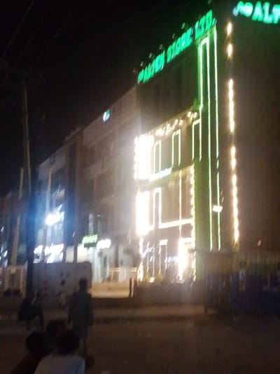The image depicts a nighttime street scene in Babban Giji, Nigeria. The central focus is a multi-story commercial building, identified by prominent green neon signage on its upper facade reading "ALPEN STORE LTD." and "ALFA." This building is extensively illuminated with bright white and green LED strip lights outlining its architectural features and window frames. To the left, a less illuminated commercial building is visible. In the foreground, a dark paved surface suggests a street or sidewalk. At least four indistinct human figures are present: three in the immediate foreground with their backs to the camera, and one further back to the left, all appearing as pedestrians. A utility pole is visible on the far left. On the far right, a blue and white structure is partially discernable. The scene is characterized by the strong artificial lighting from the primary building against an otherwise dark night sky.