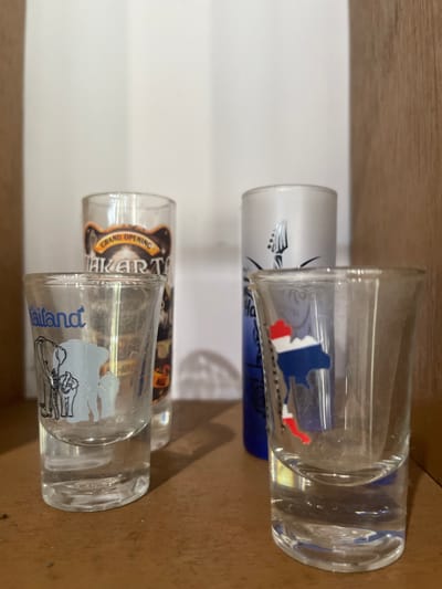 The image displays four shot glasses arranged on a wooden shelf against a white background. The shot glasses are of varying designs. The leftmost glass features a graphic of elephants and the word "Thailand" in blue. The second glass from the left is taller and features text that appears to be "JAKARTA" with "GRAND OPENING" above it, suggesting it is a souvenir from Jakarta. The third glass from the left is a frosted white glass with a graphic of a guitar. The rightmost glass has a graphic depicting a map of an area with red, white, and blue colors, possibly representing a flag or region. The shot glasses are positioned in a way that some are in focus while others are slightly blurred due to depth of field. The lighting suggests an indoor setting, possibly a shelf in a home or display cabinet. There are no people or specific actions depicted. The location is identified as Julicotmeurak, Indonesia, though this is not visually confirmed within the image.