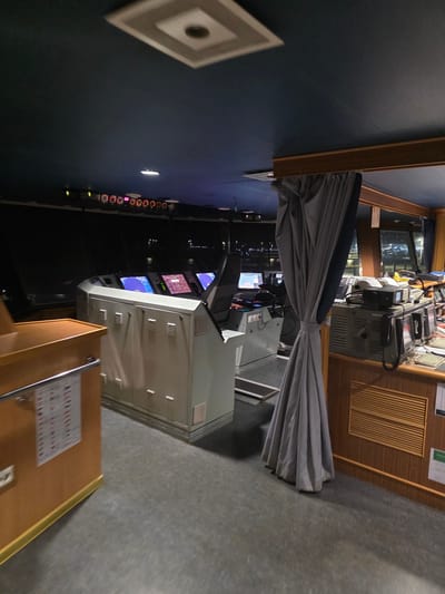 The image depicts the interior of a ship's bridge, likely at night, given the illuminated screens and distant city lights visible through the windows. The bridge is equipped with multiple electronic navigation displays, steering controls, and communication equipment. A large grey curtain hangs prominently in the foreground, partially obscuring the view of the control panels. To the left, a wooden console with charts and signage is visible. The overall impression is one of a functional, modern maritime control center. The presence of specific city lights and the layout suggest this could be the bridge of a ferry or cargo ship docked in the port of Brindisi, Italy.